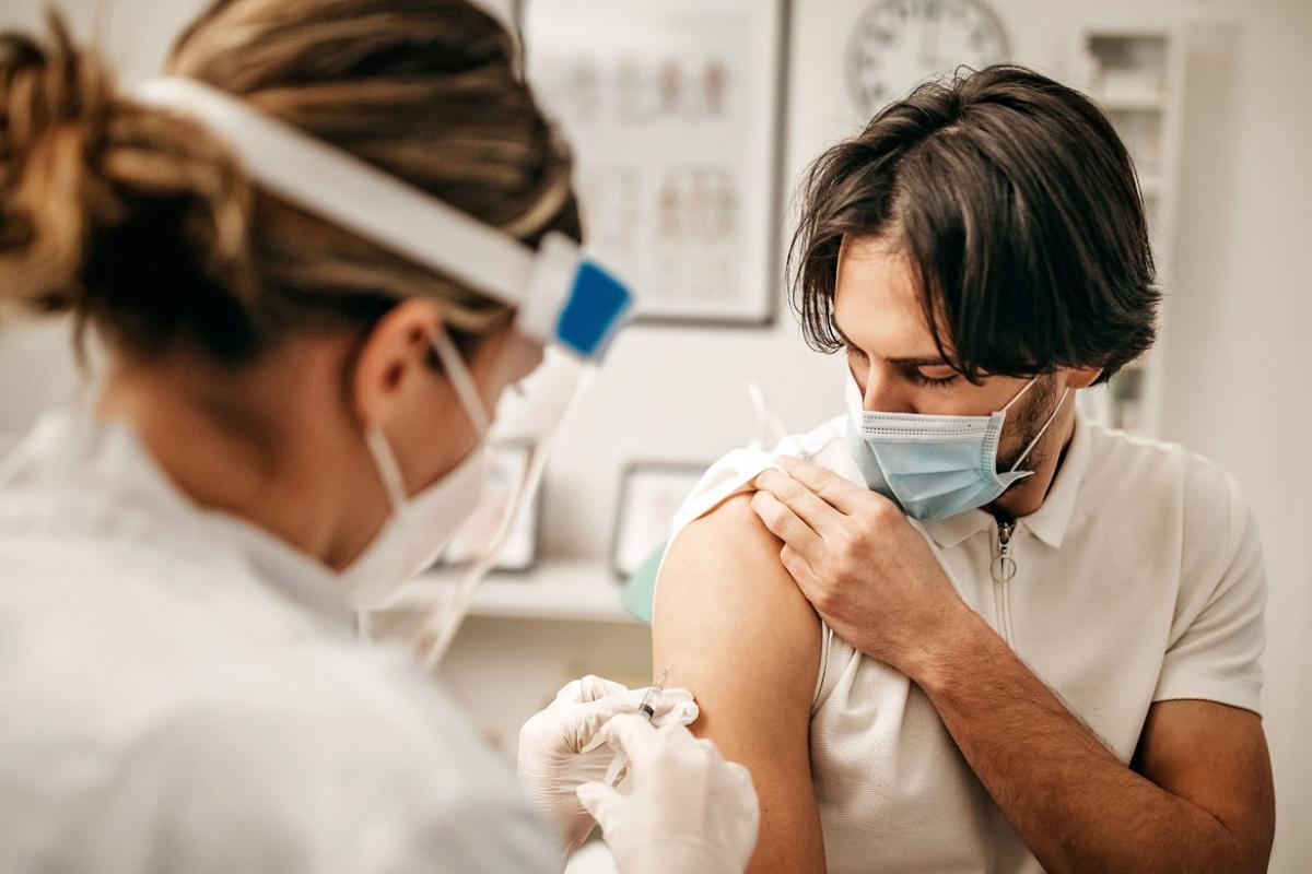 Health professional with PPE administering vaccine to right arm of patient wearing mask.