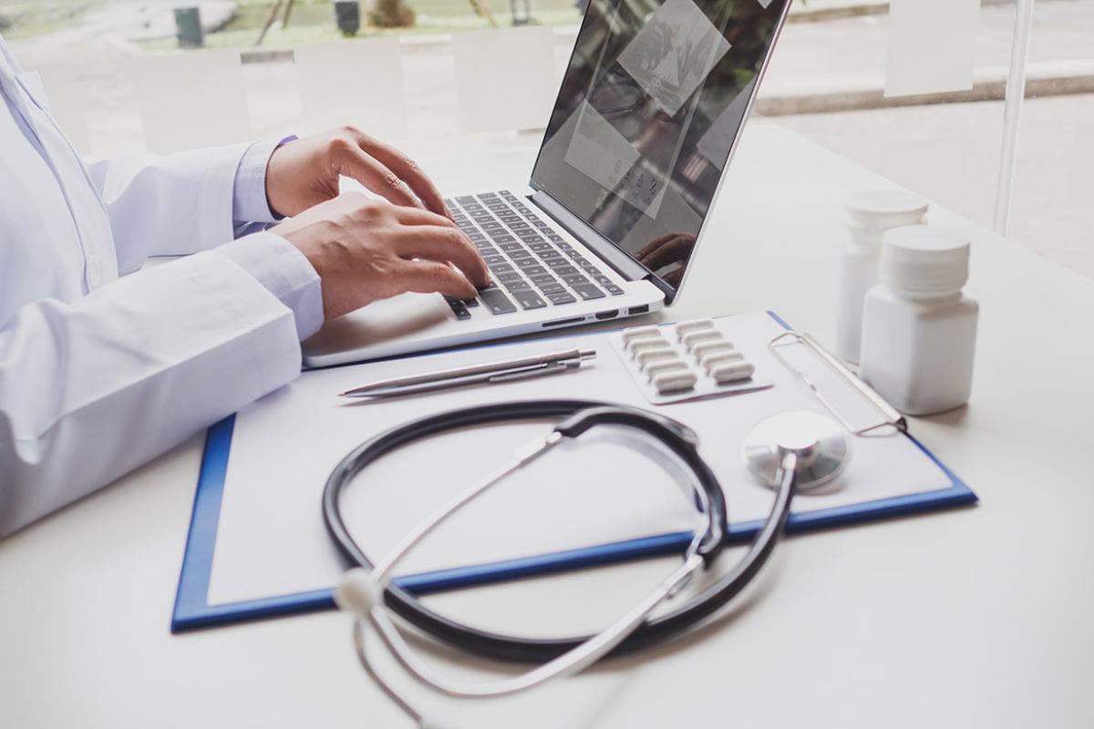 Tight shot of physician typing on a laptop keyboard, with a clipboard, stethoscope, pen, pills and prescription bottles to the side