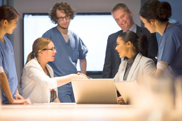 Two physicians shake hands over a laptop while 4 other physicians look on.